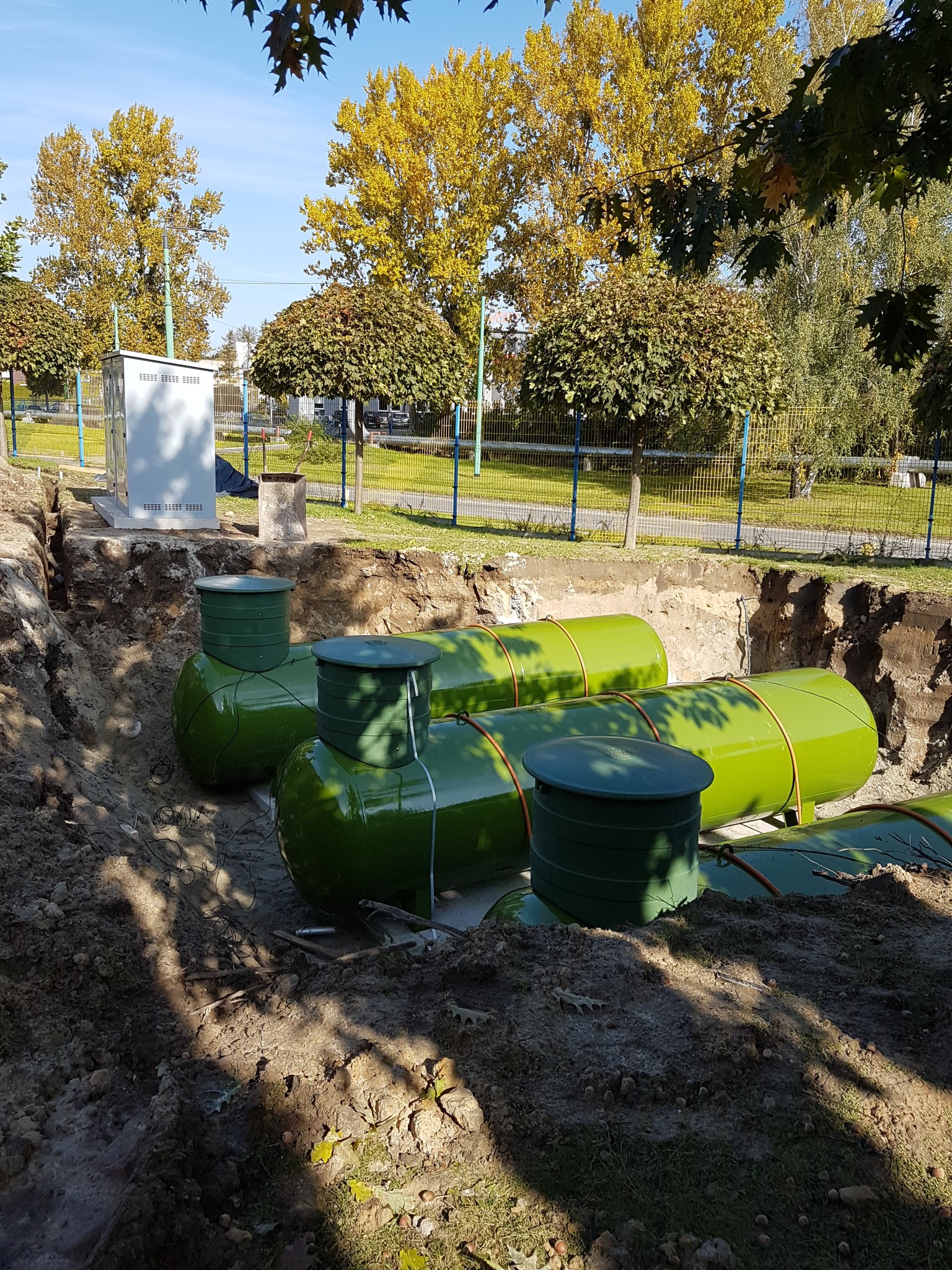 Zielone zbiorniki gazowe zakopane w wykopie ziemnym, widoczne elementy instalacji w terenie zabudowanym.