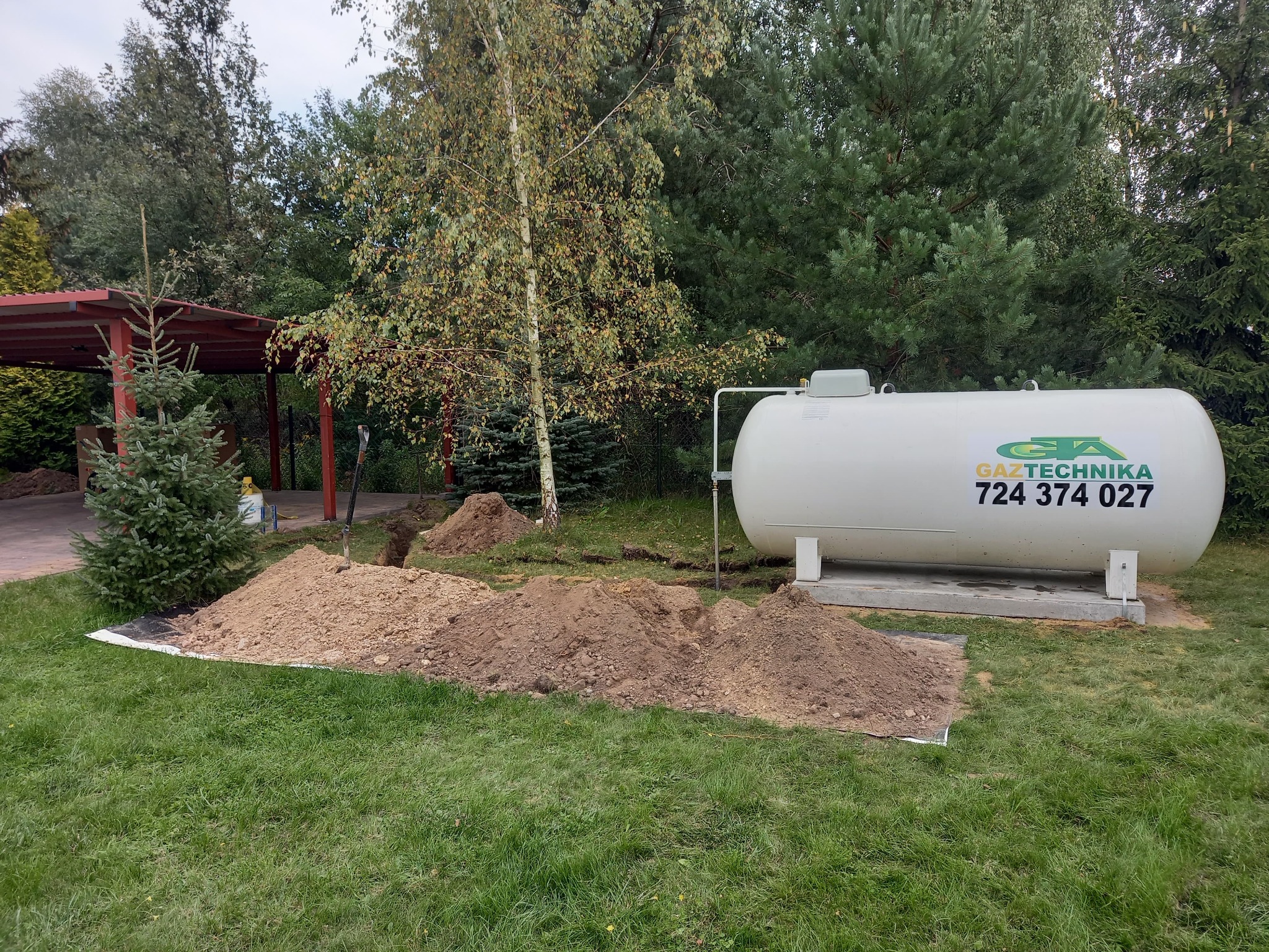 Biały zbiornik na gaz płynny z zielonym logo firmy Gaztechnika i numerem telefonu 724 374 027, ustawiony na betonowej podstawie na trawniku, obok wykopu i pryzmy ziemi, w tle drzewa i altana.