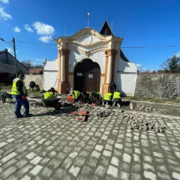 Grupa pracowników w odblaskowych kamizelkach układa kostkę granitową na placu przed zabytkową kapliczką z białymi kolumnami i zdobieniami, widoczne narzędzia i taczki.