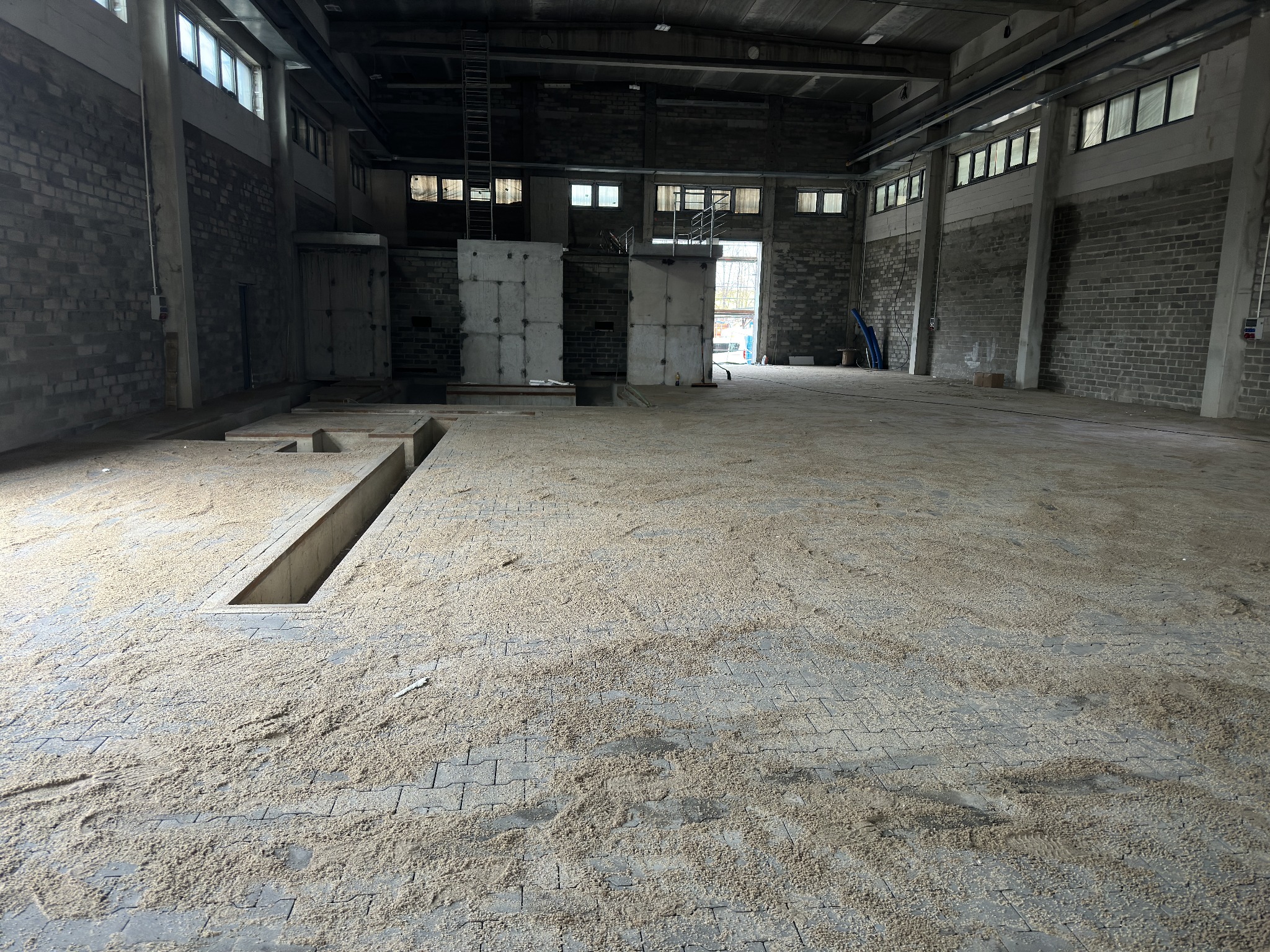 Rozległa hala z szarą kostką brukową częściowo zasypaną piaskiem, widoczne kanały rewizyjne w podłodze, surowe ściany z cegły i betonowe filary, w tle okna i metalowa konstrukcja.