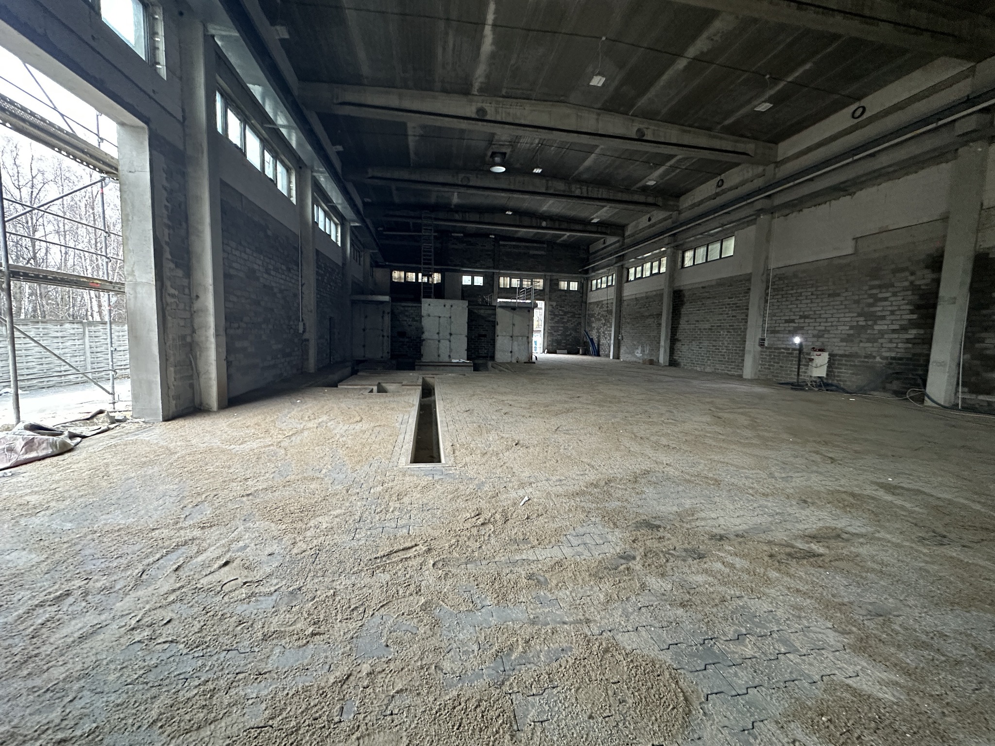 Wnętrze hali przemysłowej z częściowo ułożoną kostką brukową, pokrytą warstwą piasku, widoczny kanał odwadniający w posadzce, surowe ściany z cegły i betonowe filary.