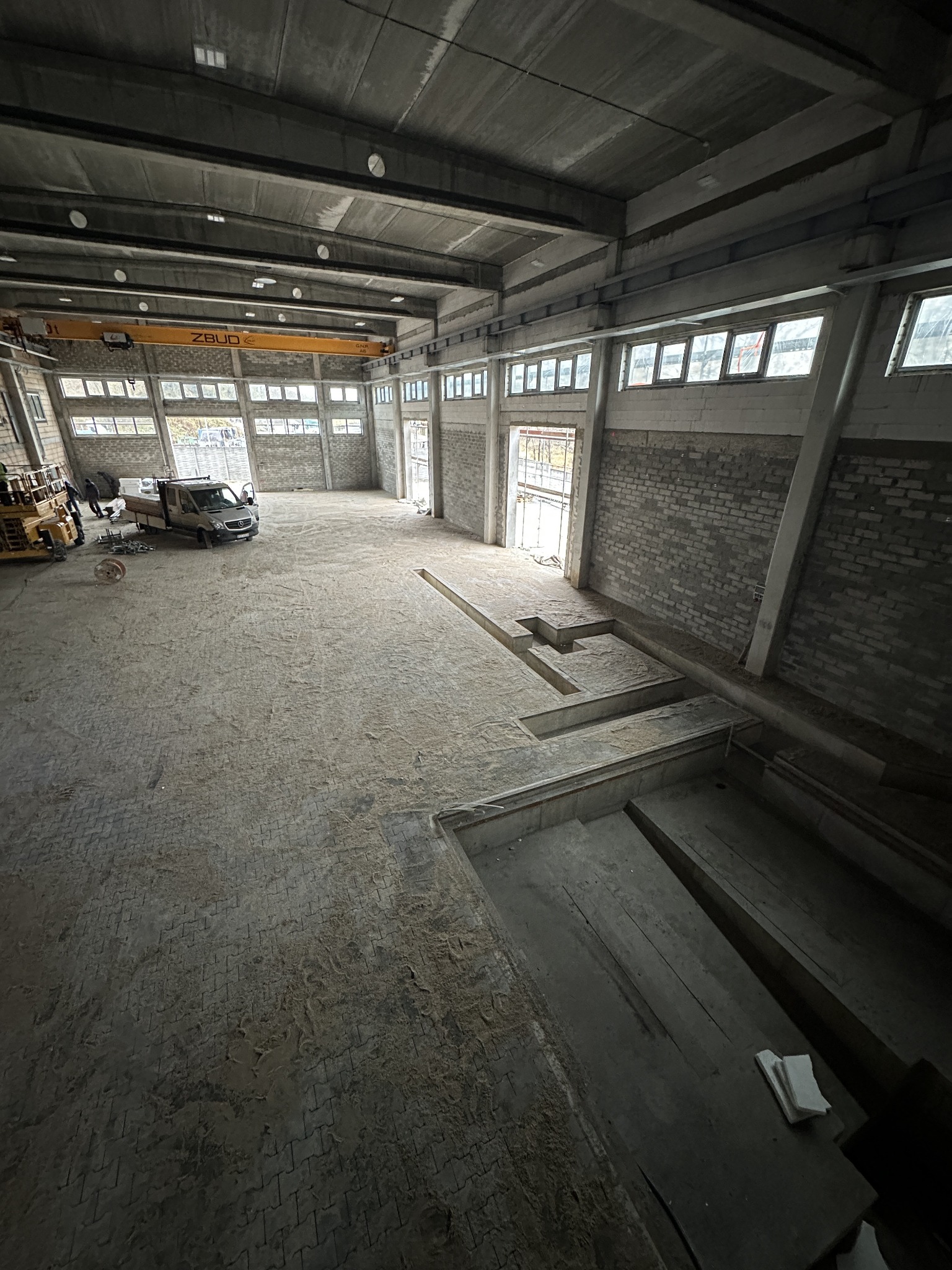 Wnętrze hali z częściowo ułożoną kostką brukową, widoczne wykopane fragmenty podłoża, pojazd dostawczy i maszyny budowlane w tle, surowy betonowy strop z suwnicą.