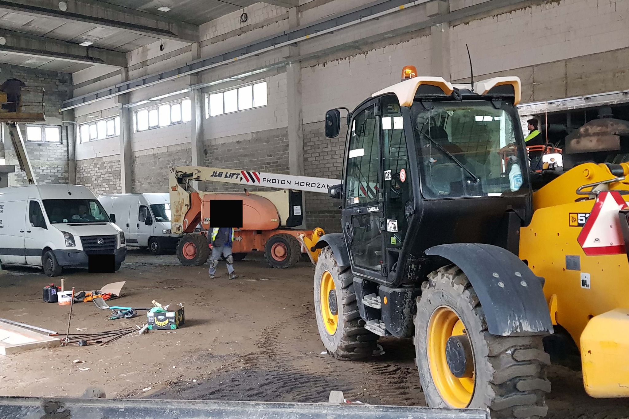 Wnętrze hali z zaparkowanymi samochodami dostawczymi, podnośnikami koszowymi i ładowarką teleskopową JCB 531-70 Loadall, widoczne narzędzia i materiały budowlane.