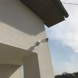 Zewnętrzna kamera monitoringu Dahua zamontowana na rogu budynku z szarą elewacją, widok z dołu na tle błękitnego nieba z chmurami.