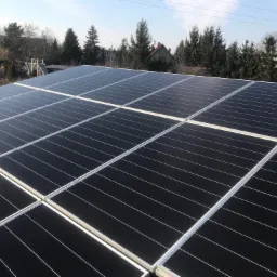 Panele fotowoltaiczne zamontowane na dachu domu jednorodzinnego, widoczne z bliska pod kątem, z domami i drzewami w tle pod błękitnym niebem.