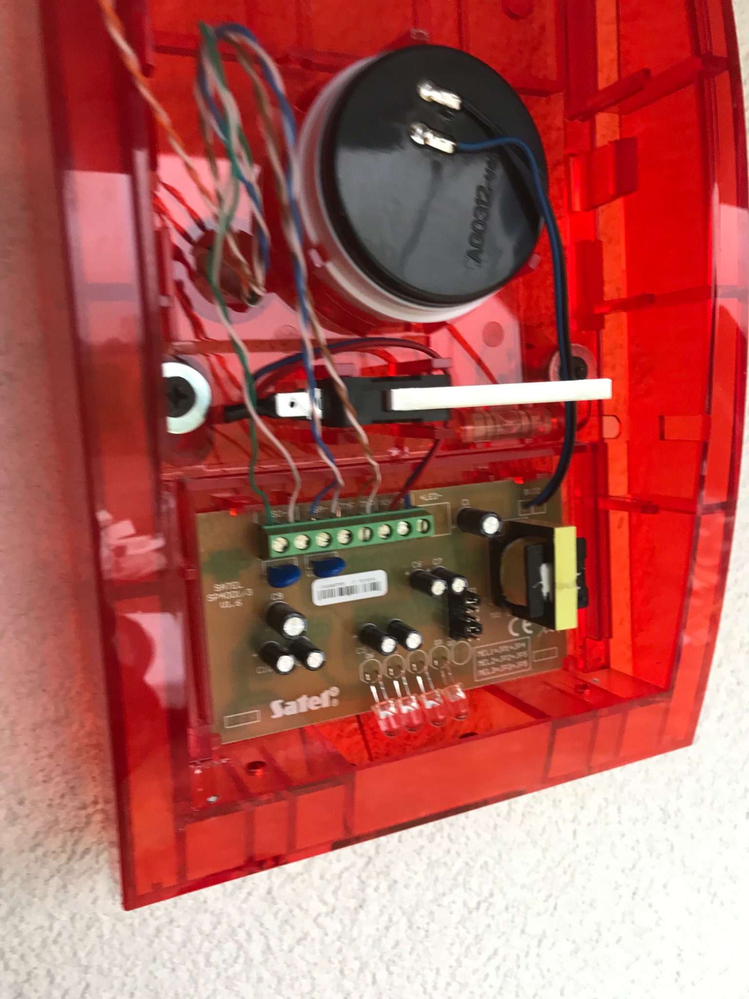 Wnętrze czerwonej obudowy alarmu z widoczną płytką drukowaną, głośnikiem i przewodami podłączonymi do złączek.