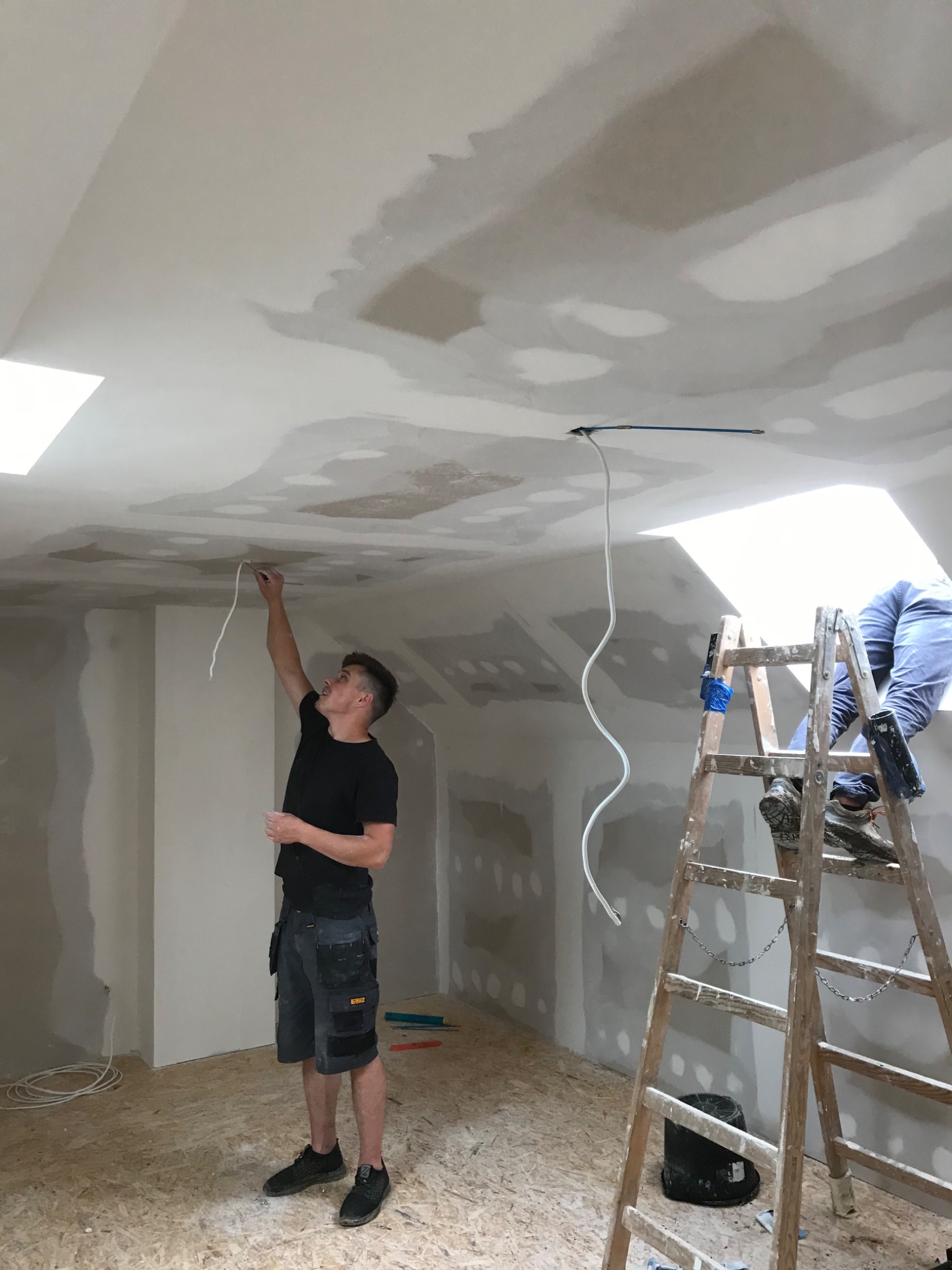 Elektryk przeciąga przewód elektryczny w suficie podwieszanym w trakcie remontu poddasza. Widoczna drabina, narzędzia i fragment instalacji.