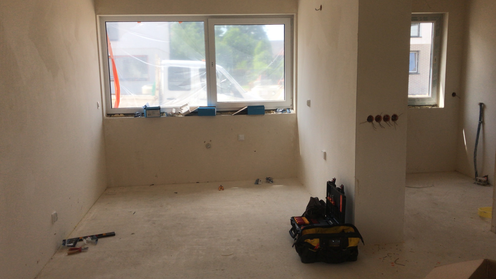 Wnętrze w stanie deweloperskim z widocznymi puszkami elektrycznymi na ścianie, skrzynką narzędziową na podłodze i oknami zabezpieczonymi folią.