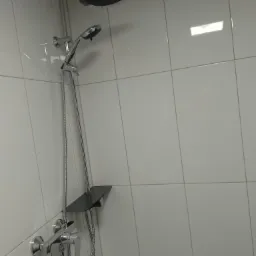 Bateria prysznicowa z deszczownicą i słuchawką, zamontowana na ścianie wyłożonej białymi, błyszczącymi płytkami ceramicznymi; widoczna półka na kosmetyki.