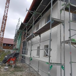 Solid Renovate - Elewacja budynku w trakcie prac wykończeniowych, rusztowania, maszyna do nakładania tynku, widoczny fragment żurawia.  Szara, pochmurna pogoda.