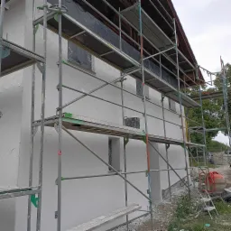 Elewacja budynku w trakcie prac wykończeniowych, widoczne rusztowania, ślady zaprawy i mieszalnik. Okna zabezpieczone folią.