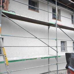 Solid Renovate - Elewacja budynku w trakcie prac wykończeniowych, widoczne rusztowanie, okna zabezpieczone folią, świeżo nałożona struktura, fragment drabiny i maszyny do nakładania tynku.