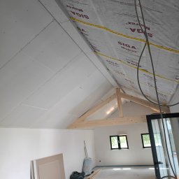 Solid Renovate - Poddasze w trakcie wykańczania: białe płyty gipsowe na skośnym suficie, drewniana konstrukcja więźby dachowej, okna w ścianie szczytowej, materiały budowlane na podłodze.