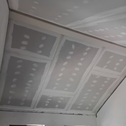 Solid Renovate - Sufit podwieszany z płyt gipsowo-kartonowych, widoczne łączenia i miejsca po wkrętach, przygotowany do dalszych prac wykończeniowych. W tle okno i poziomica.