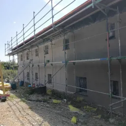 Budynek w trakcie prac wykończeniowych. Na elewacji widoczne rusztowania, ściany pokryte szarą warstwą, okna zabezpieczone folią. Na ziemi leżą worki z materiałami.