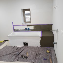 Solid Renovate - Biała wanna obudowywana szarą zaprawą, zabezpieczona fioletową taśmą. Na podłodze rozłożona mata ochronna i narzędzia glazurnicze. Widoczne przyłącza wodne.