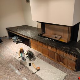 Solid Renovate - Montaż obudowy kominka z kamienia. Widoczne narzędzia, płyty kamienne i fragment wkładu kominkowego. Prace wykończeniowe w trakcie realizacji.