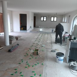 Solid Renovate - Układanie płytek podłogowych w jasnym wnętrzu. Widoczne narzędzia, płytki, system poziomowania i pracownik w trakcie pracy. Duża przestrzeń z oknami i kolumną.