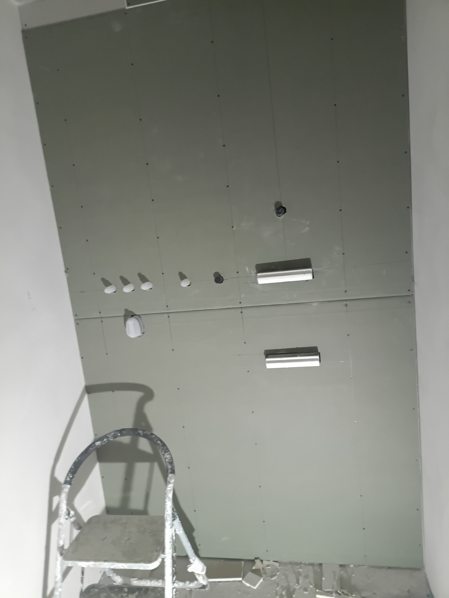 Montaż płyt gipsowo-kartonowych na ścianie z widocznymi otworami na instalacje i drabinką malarską w rogu pomieszczenia. Widoczny etap prac wykończeniowych.