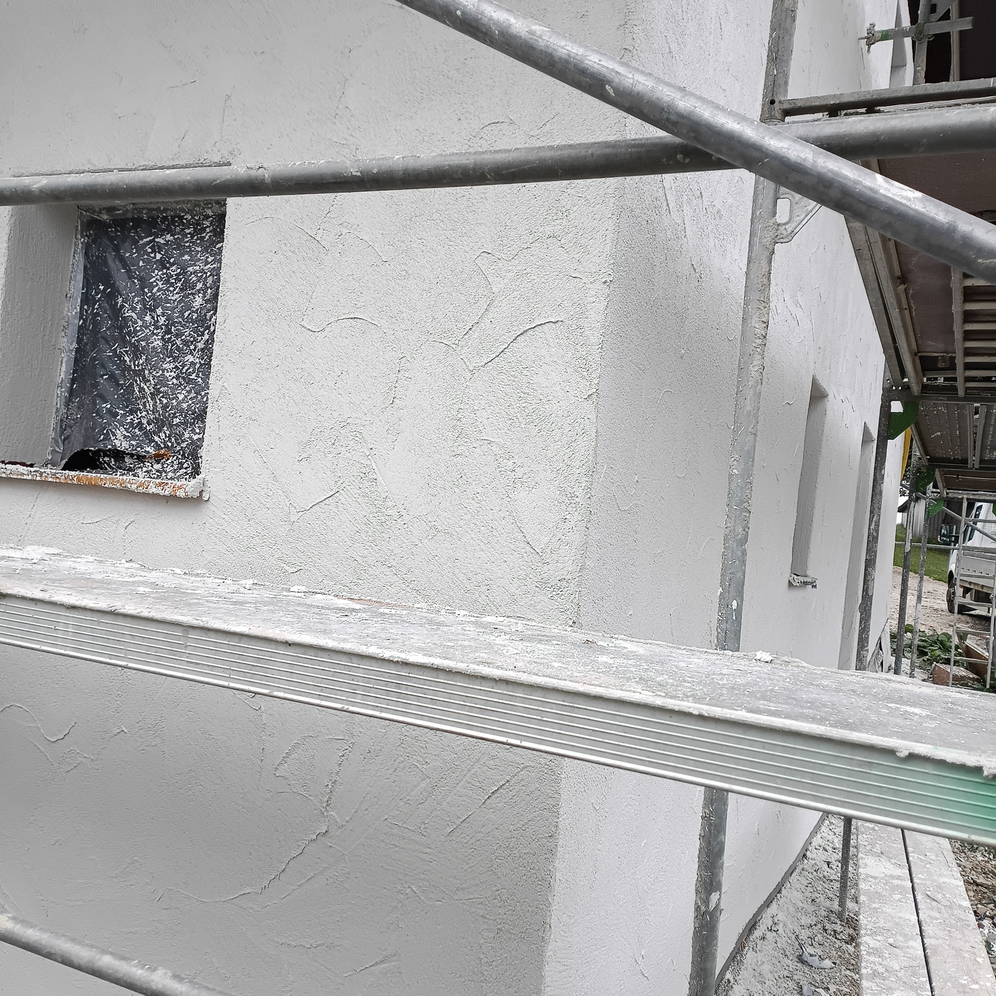 Ściana budynku w trakcie prac wykończeniowych z nałożonym tynkiem strukturalnym, zabezpieczone okno folią i rusztowanie. Widok z bliska.