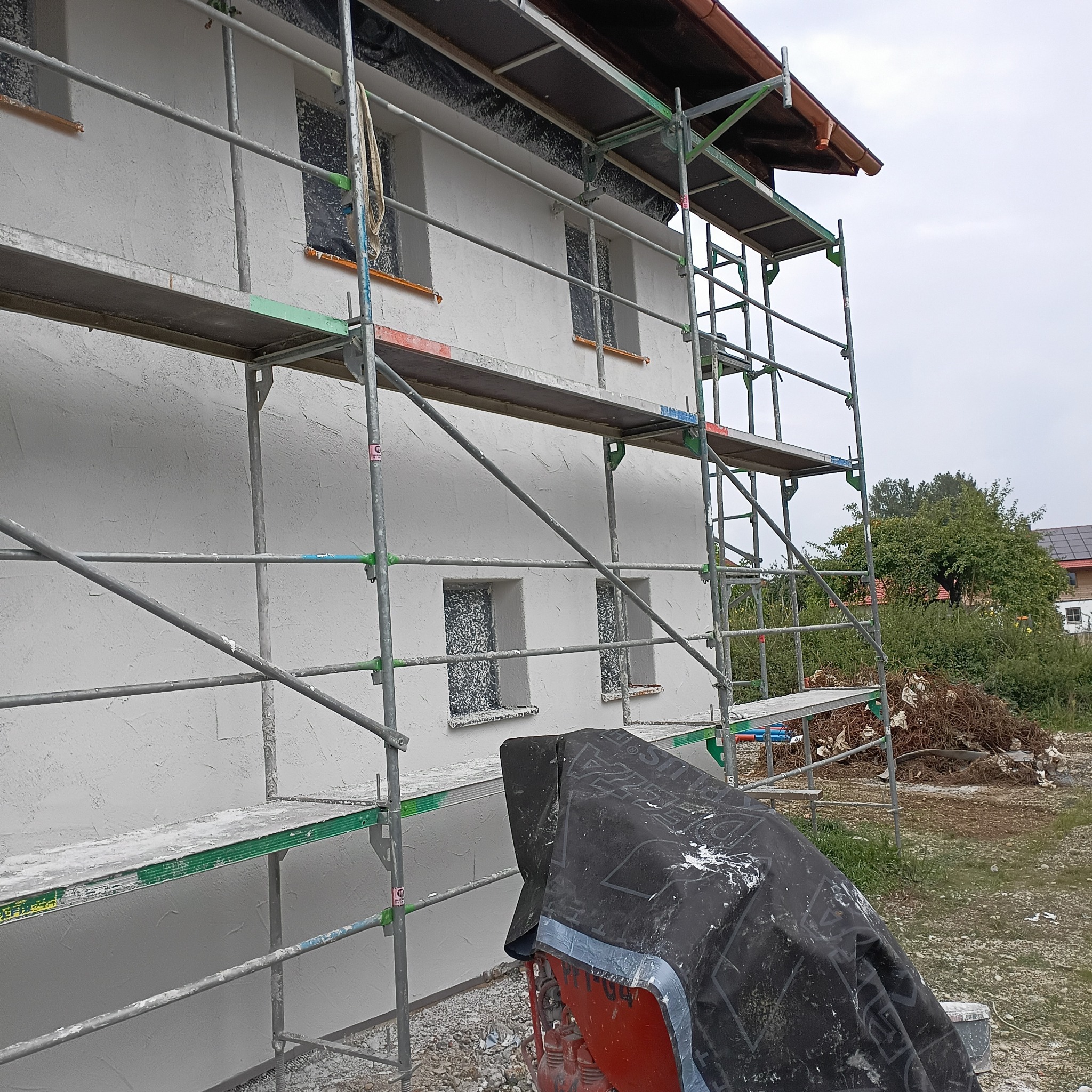 Elewacja budynku po nałożeniu białego tynku, widoczne rusztowania i zabezpieczone okna w Pelplinie. Teksturowana powierzchnia ściany.