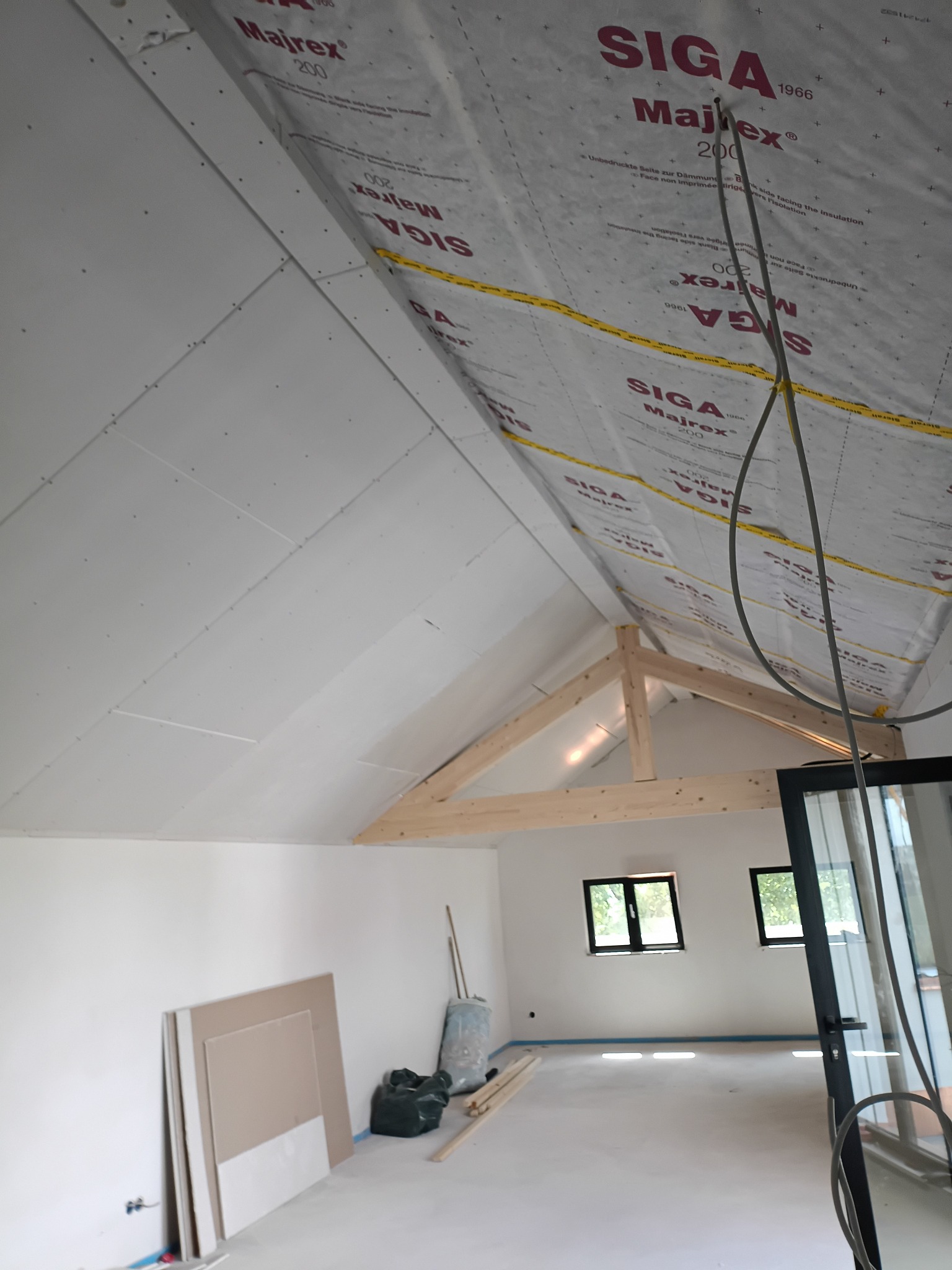 Poddasze w trakcie wykańczania: białe płyty gipsowe na skośnym suficie, drewniana konstrukcja więźby dachowej, okna w ścianie szczytowej, materiały budowlane na podłodze.