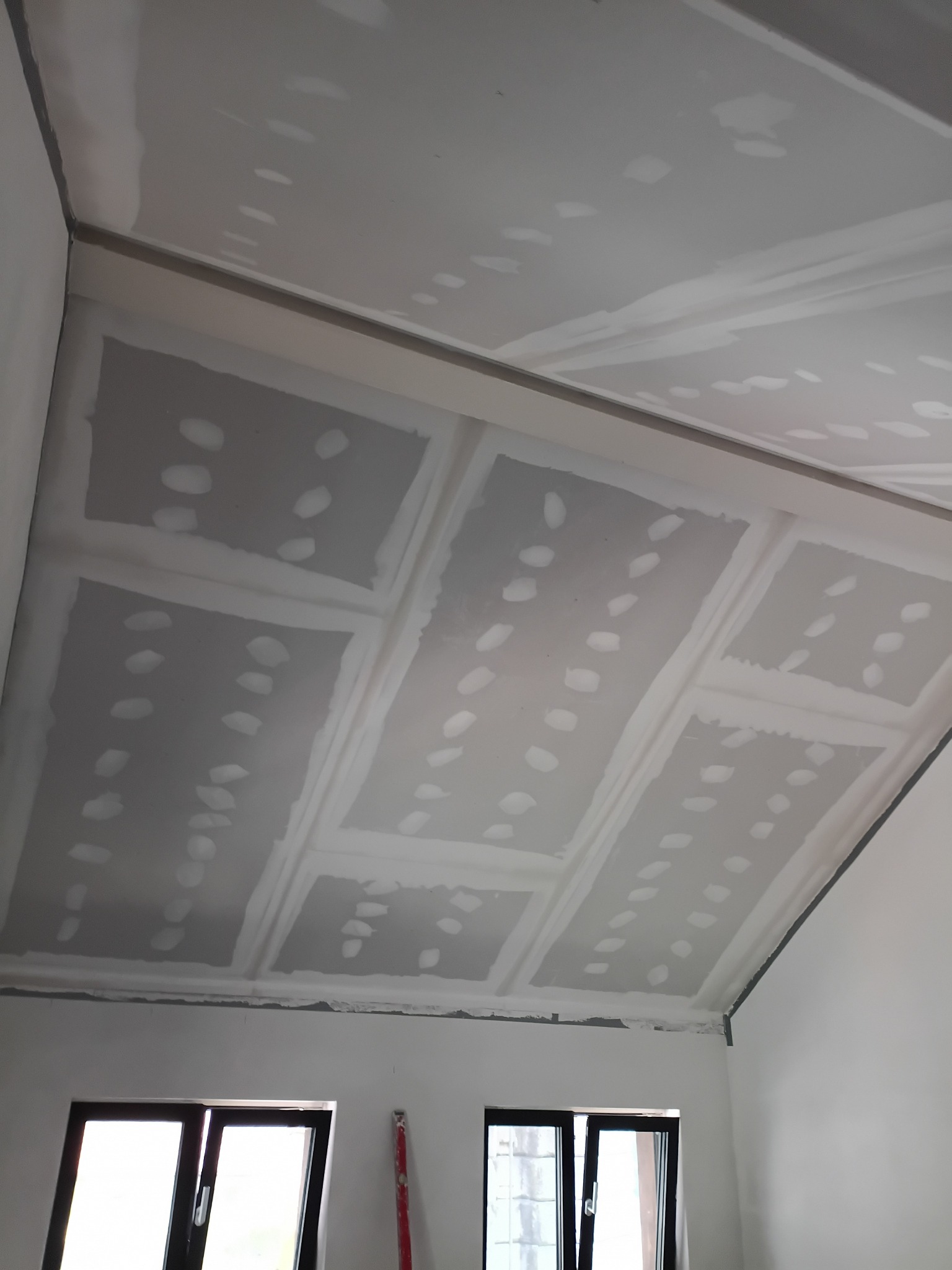 Sufit podwieszany z płyt gipsowo-kartonowych, widoczne łączenia i miejsca po wkrętach, przygotowany do dalszych prac wykończeniowych. W tle okno i poziomica.