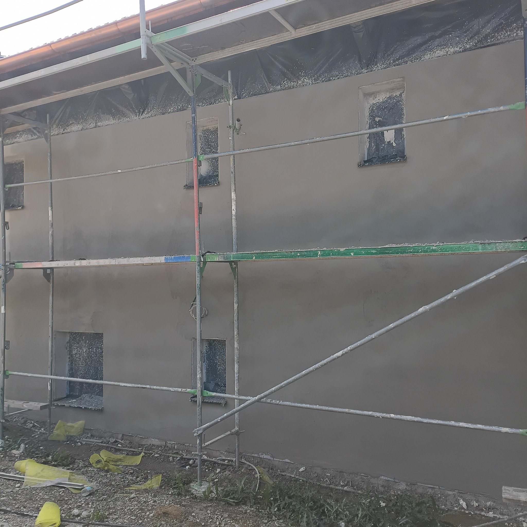 Ściana budynku w trakcie prac wykończeniowych, pokryta szarą warstwą, z widocznymi rusztowaniami i zabezpieczonymi oknami folią.