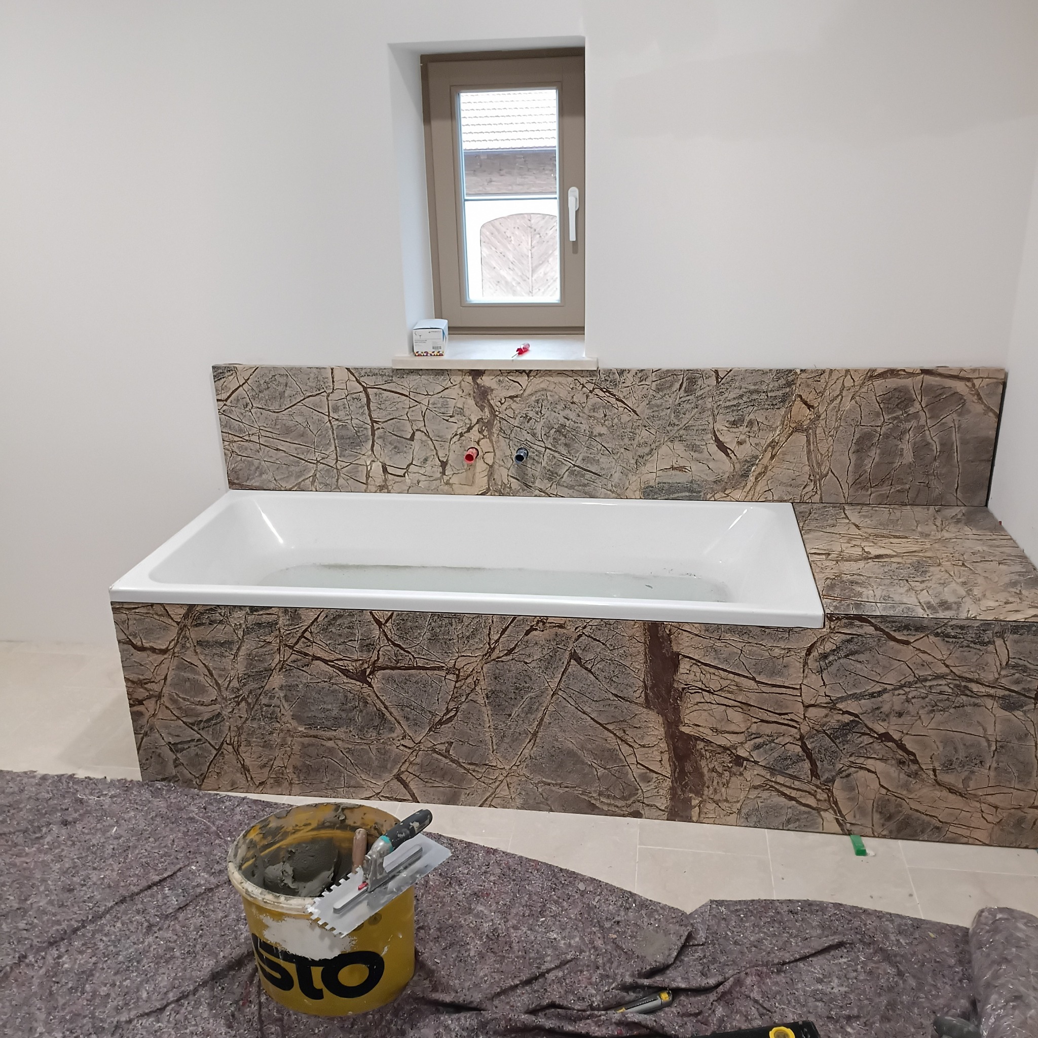 Obudowa wanny wykończona płytkami imitującymi kamień z wyraźnym wzorem żyłkowania. Widoczny fragment podłogi zabezpieczony matą ochronną i narzędzia glazurnicze.