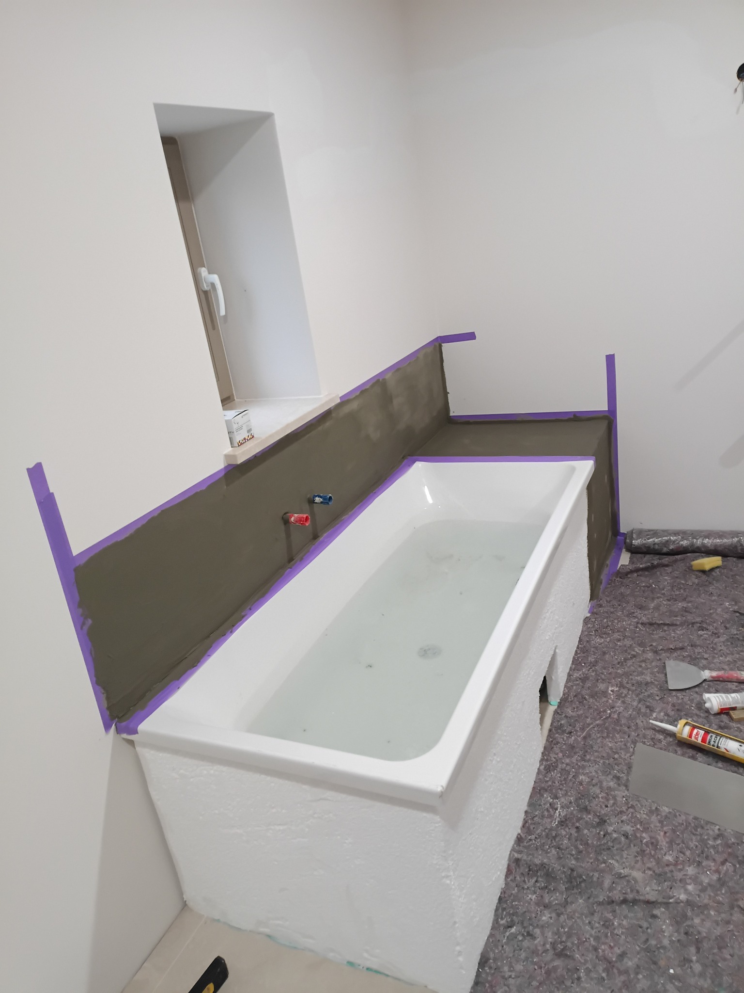 Wanna obudowana i przygotowana do wykończenia, zabezpieczona taśmą malarską. Widoczne narzędzia i materiały budowlane. Prace remontowe w łazience.