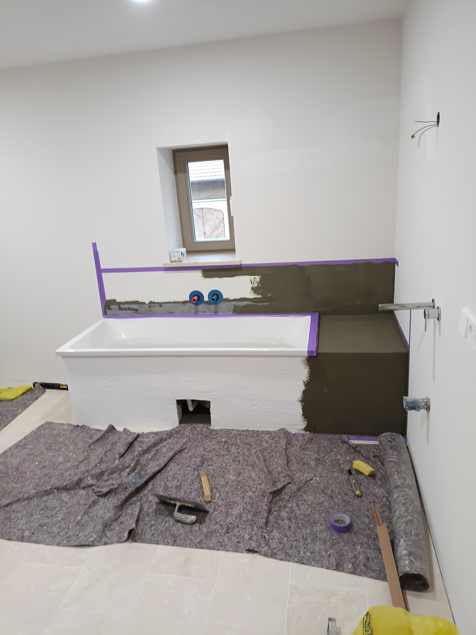Biała wanna obudowywana szarą zaprawą, zabezpieczona fioletową taśmą. Na podłodze rozłożona mata ochronna i narzędzia glazurnicze. Widoczne przyłącza wodne.