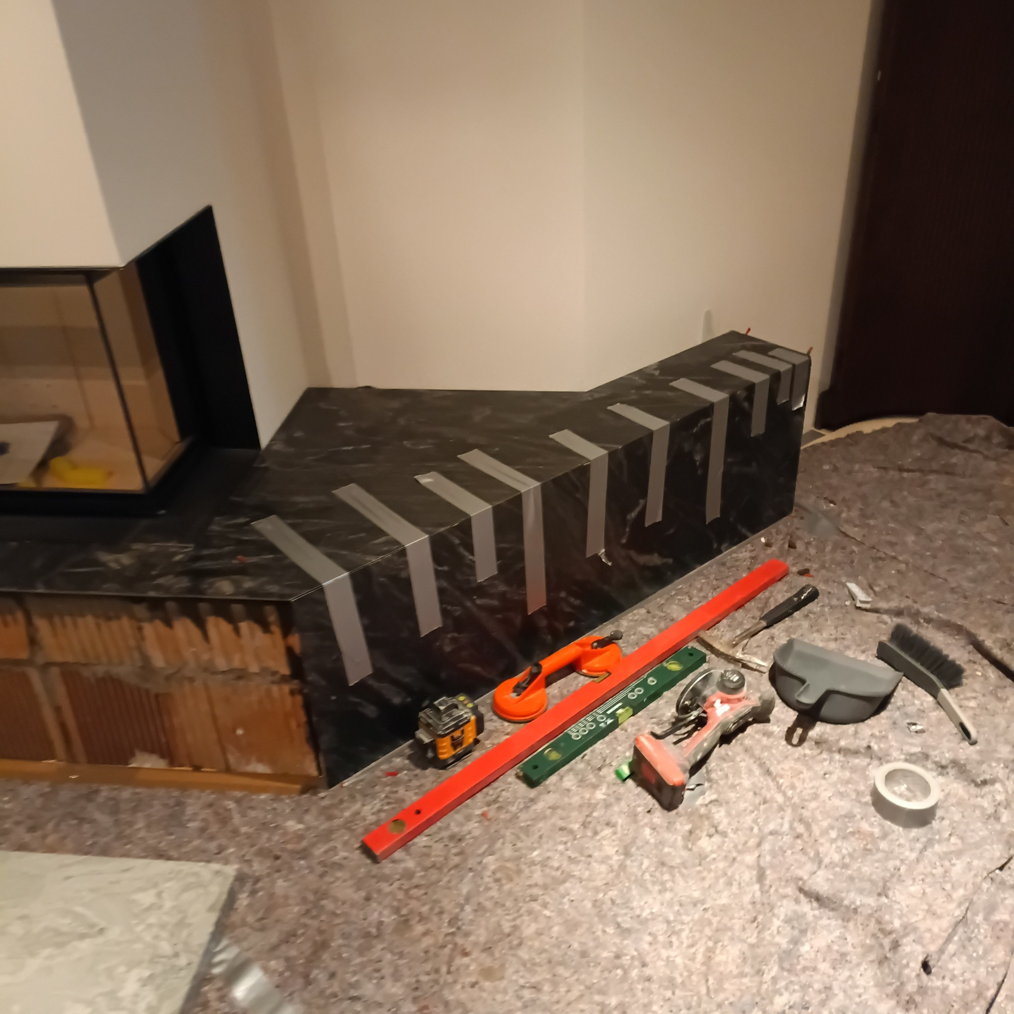 Obudowa kominka z czarnego kamienia, zabezpieczona taśmą, w trakcie montażu. Widoczne narzędzia glazurnicze: poziomica, przyssawka, laser krzyżowy.