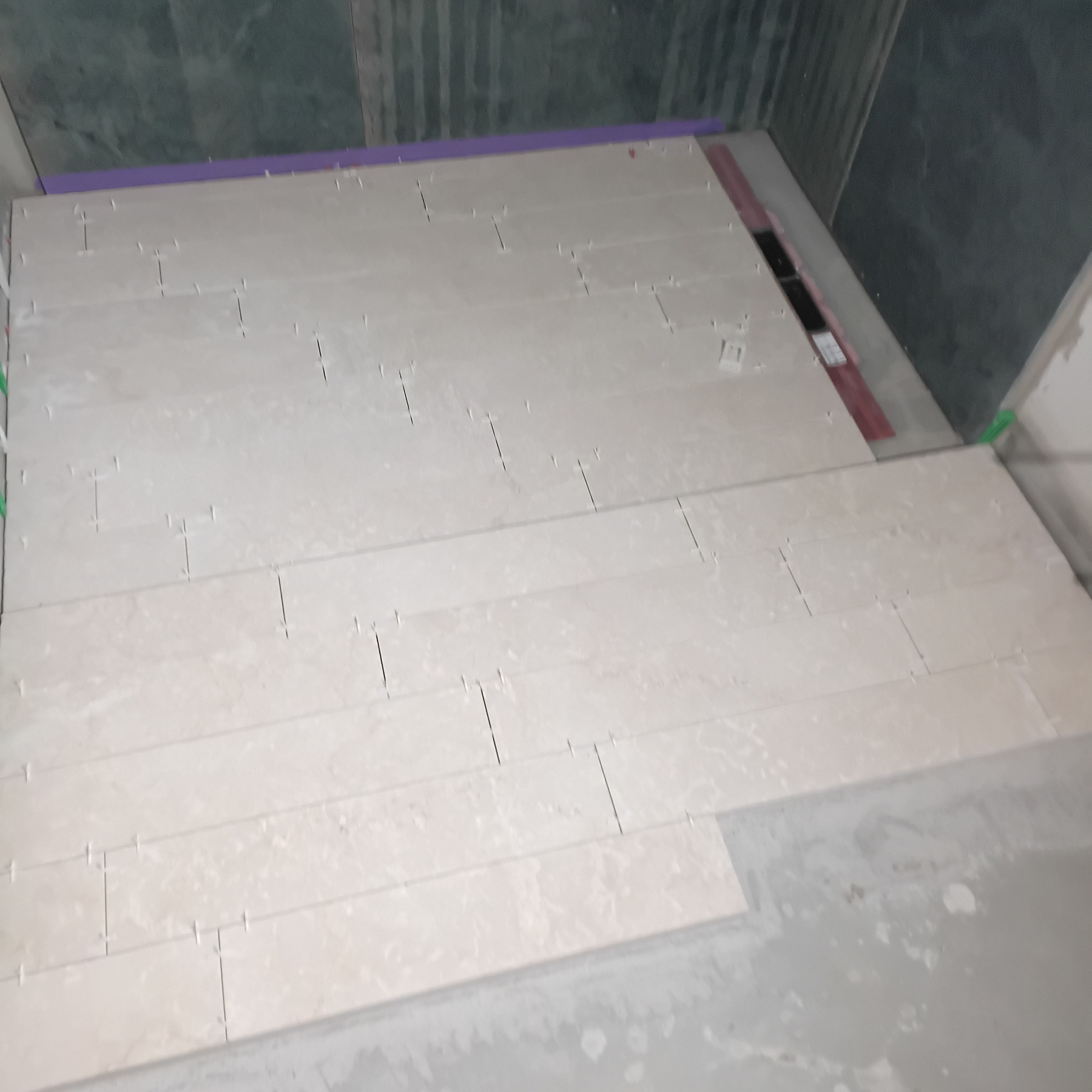 Układanie beżowych płytek podłogowych w łazience, widoczne krzyżyki dystansowe i poziomica. Częściowo ułożona podłoga, zielone ściany w tle.