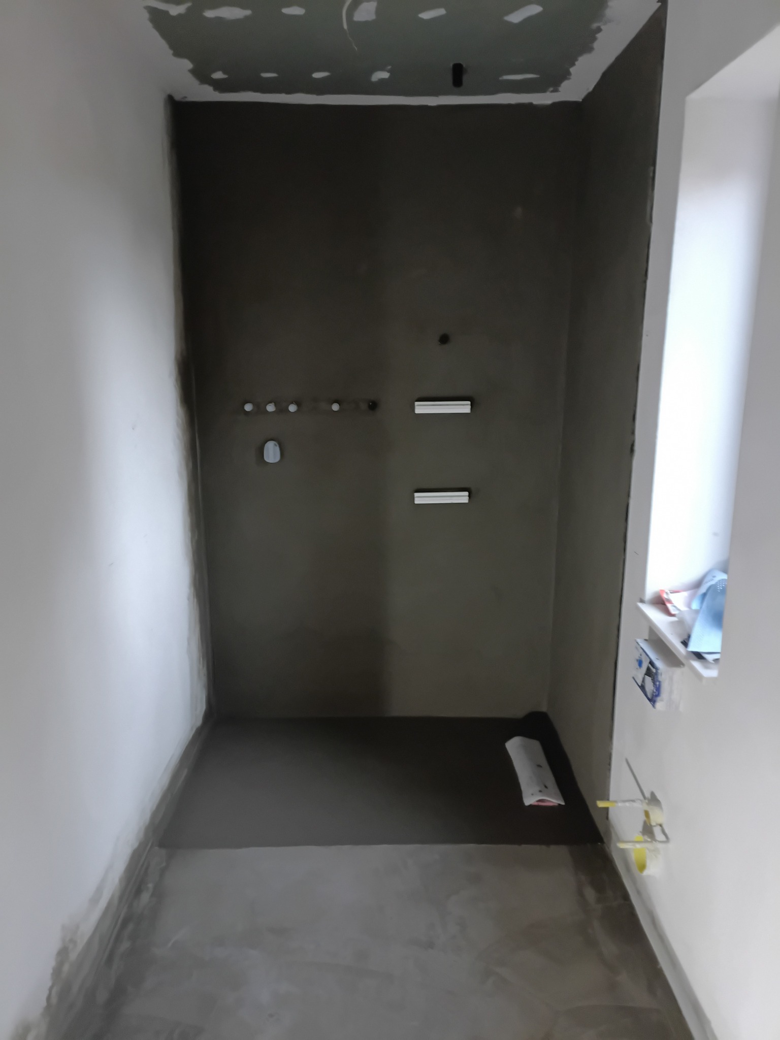 Surowa łazienka w trakcie prac: ściany z szarym tynkiem, przygotowane punkty instalacji, wylewka na podłodze. Widok na okno z akcesoriami. Minimalistyczny, nowoczesny styl.