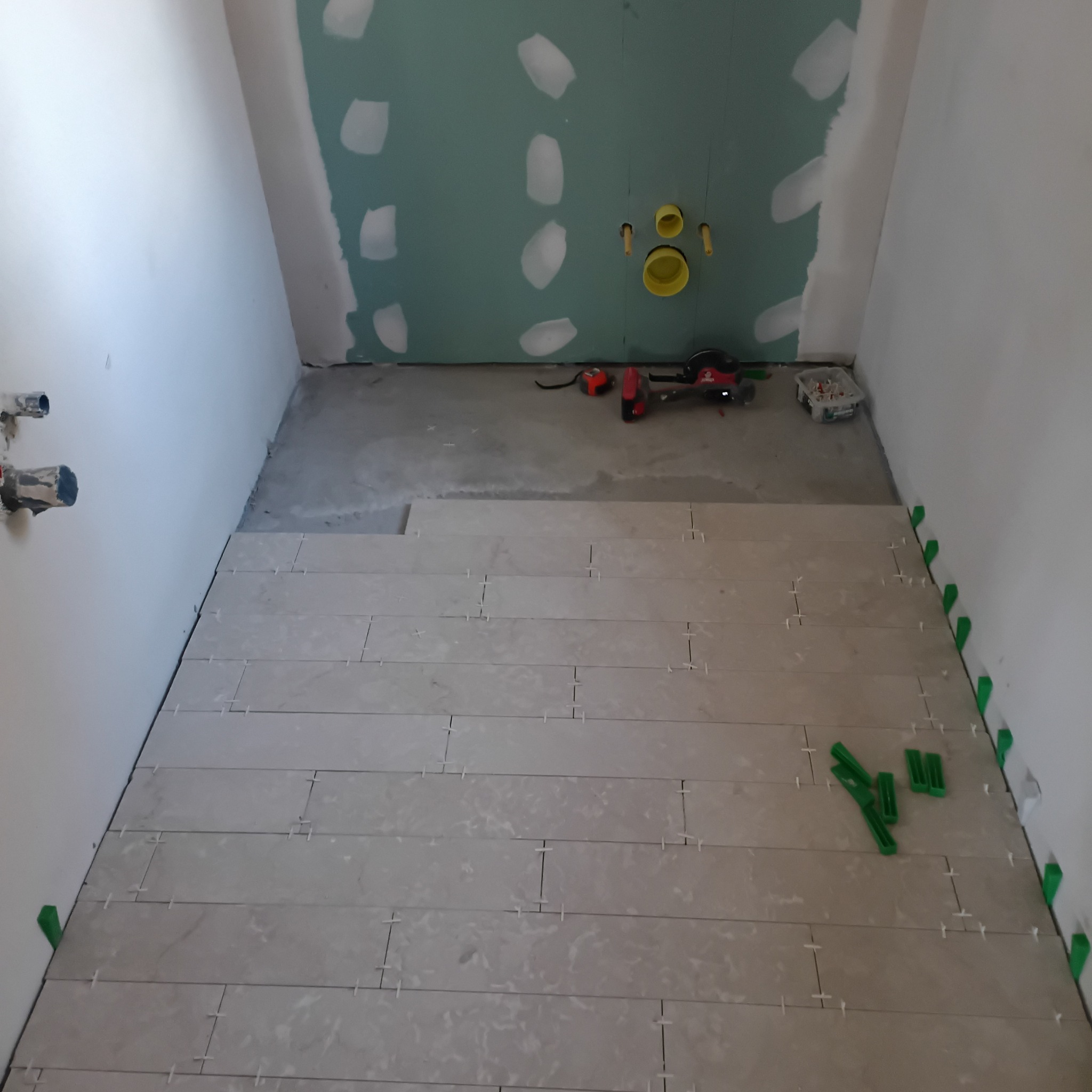 Układanie podłogi z płytek w łazience w trakcie remontu. Widoczne narzędzia, poziomica i krzyżyki dystansowe. Częściowo położone płytki i przygotowane ściany.