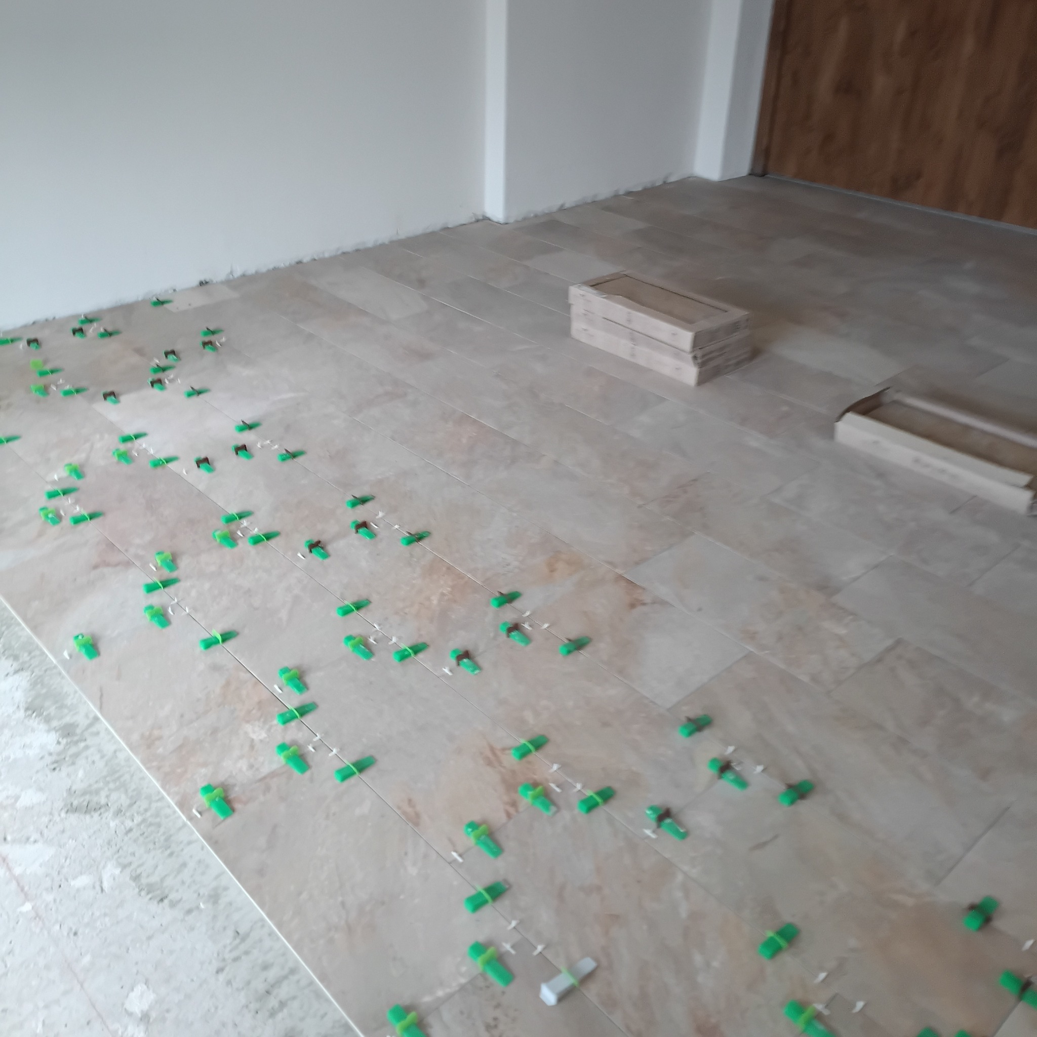 Jasne płytki podłogowe w trakcie układania, z użyciem zielonych klinów poziomujących. W tle widoczne kartony z płytkami i fragment ściany.