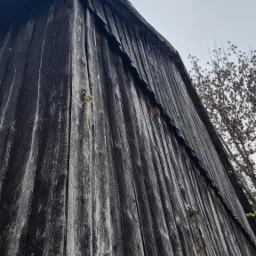 Pionowe ujęcie z dołu na starą, drewnianą konstrukcję z widocznymi śladami zużycia, sznurkiem zwisającym z desek i fragmentem drzewa w tle na pochmurnym niebie.