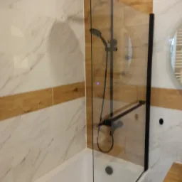 Nowoczesna łazienka z białą wanną, czarną armaturą i szklanym parawanem nawannowym w czarnej ramie, wykończona płytkami imitującymi marmur i drewno.