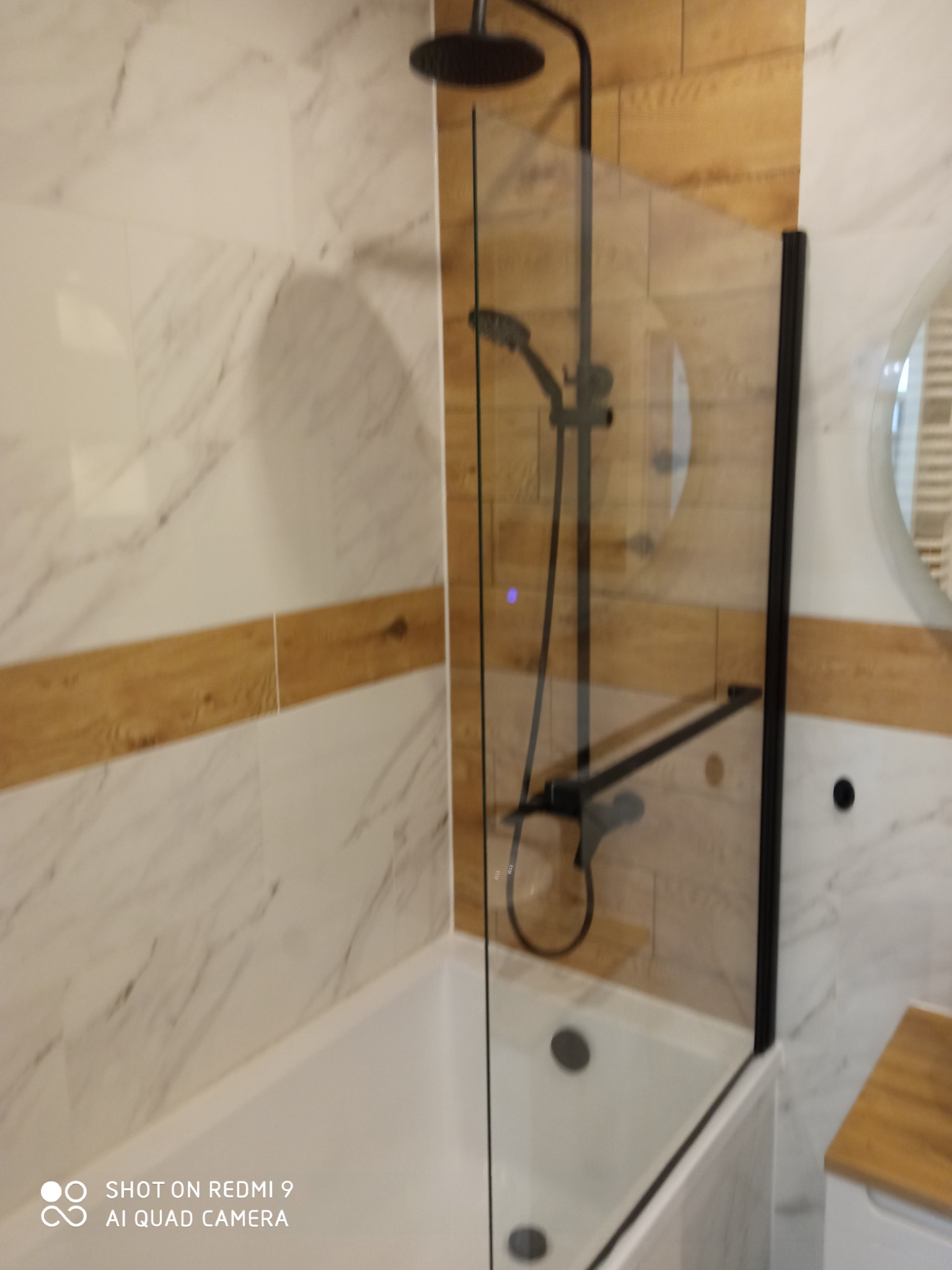 Nowoczesna łazienka z białą wanną, czarną armaturą i szklanym parawanem nawannowym w czarnej ramie, wykończona płytkami imitującymi marmur i drewno.