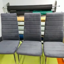 Trzy krzesła z szarą tapicerką i chromowanymi nogami ustawione na tle rolek materiałów w pracowni.