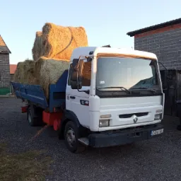 Biała ciężarówka Renault z niebieską skrzynią ładunkową wypełnioną balotami siana na żwirowym podłożu, na tle murowanego budynku.