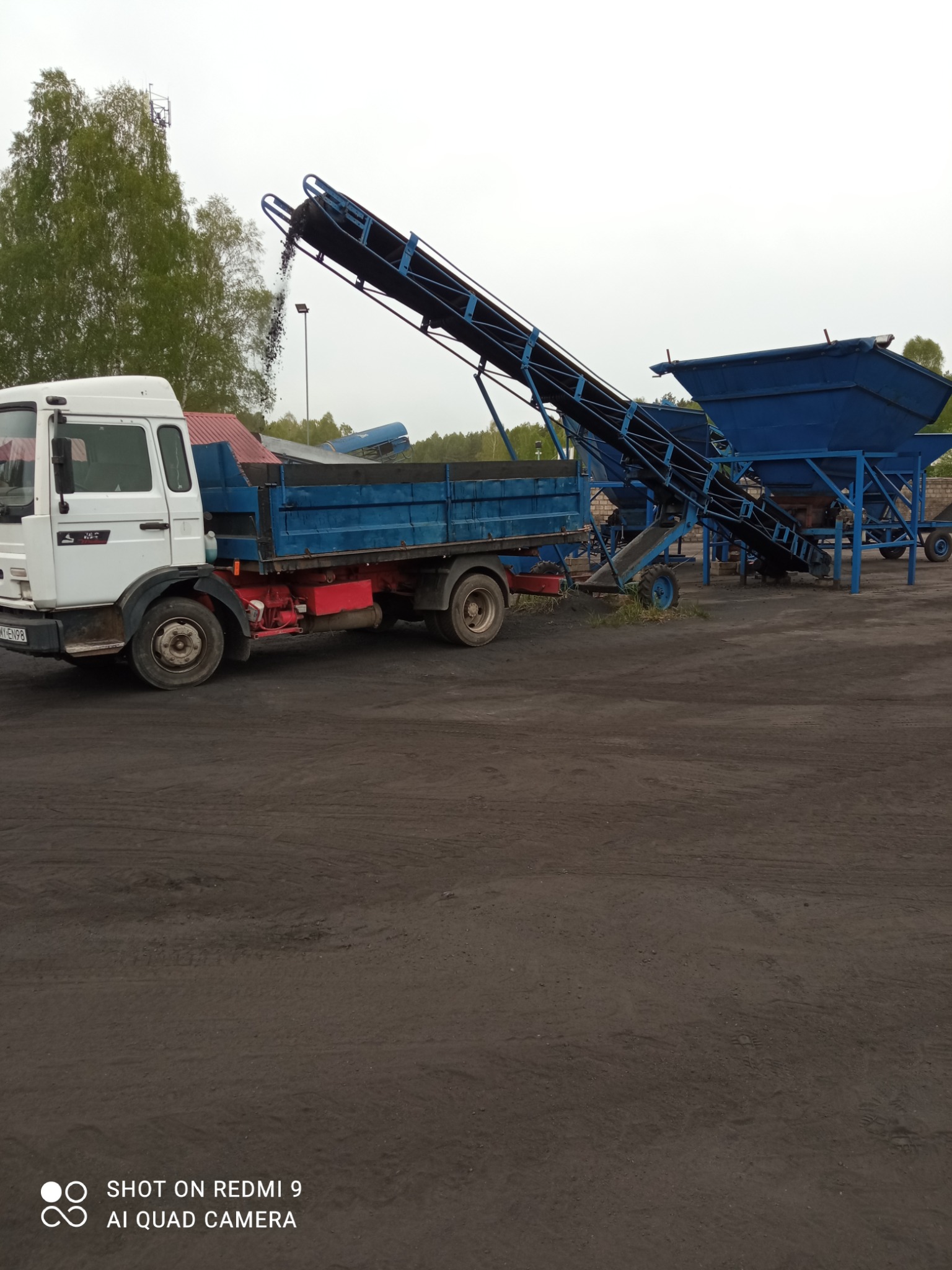 Biała ciężarówka z niebieską skrzynią ładunkową pod przenośnikiem taśmowym, który wysypuje ciemny materiał do skrzyni. W tle niebieski przesiewacz i drzewa.