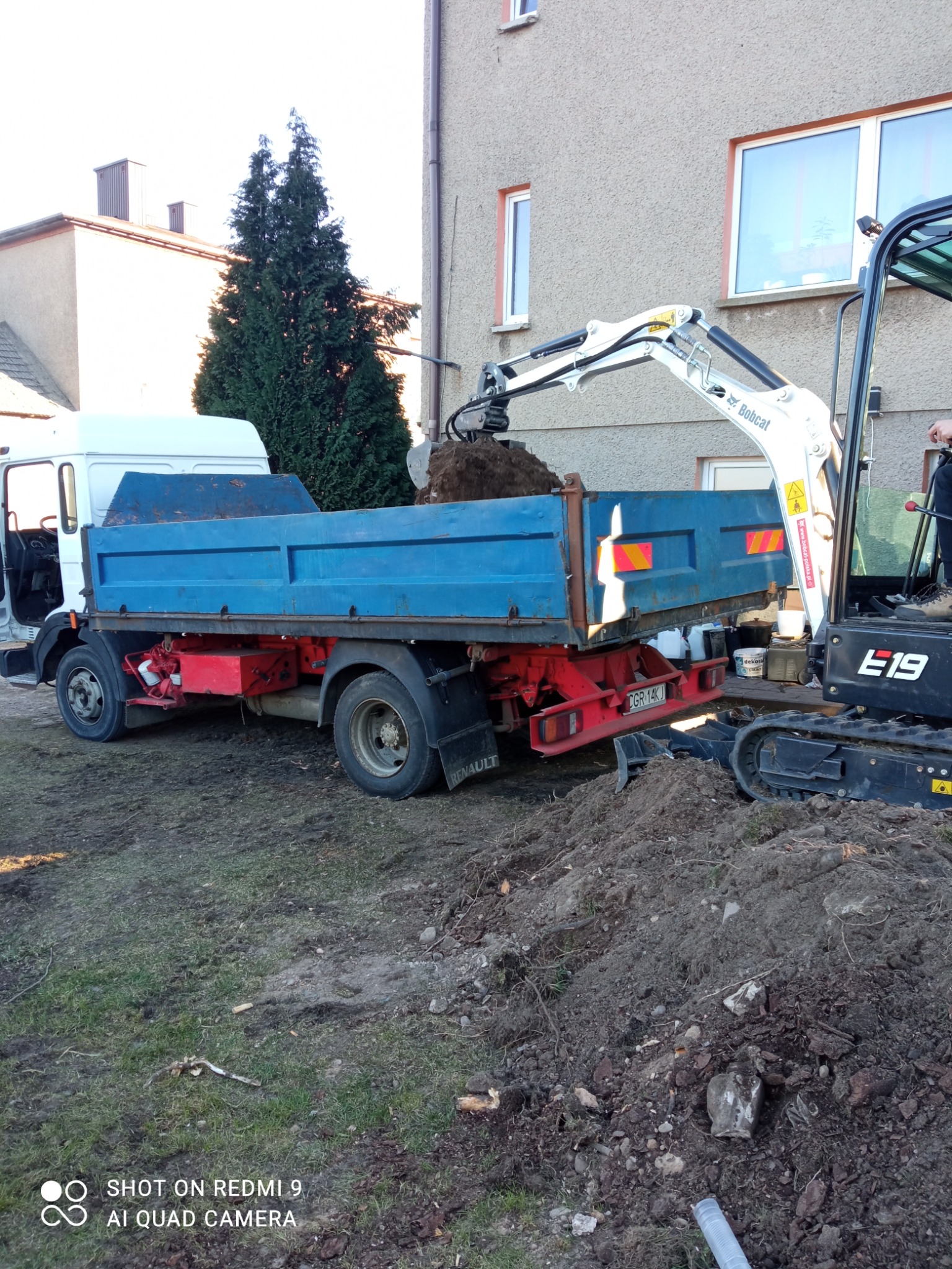 Niebieska ciężarówka Renault z otwartą kabiną ładuje ziemię z wykopu za pomocą minikoparki Bobcat E19, w tle budynek mieszkalny i iglaste drzewo.