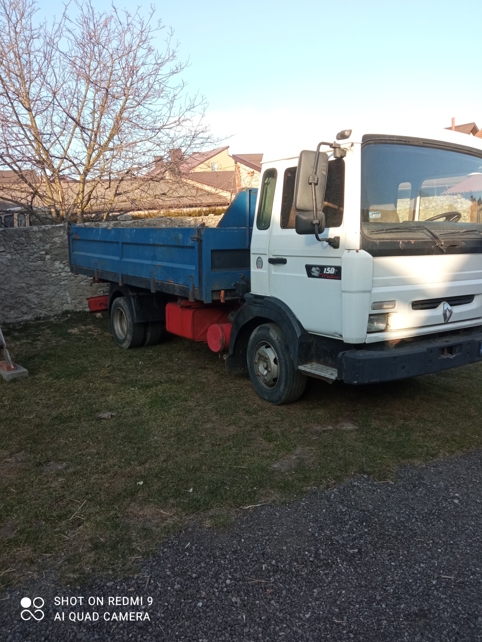 Biała ciężarówka Renault 150 z niebieską skrzynią ładunkową, stojąca na trawie obok kamiennego muru i drzewa bez liści, w tle widoczne dachy budynków.