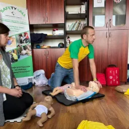 Instruktor w koszulce żółto-zielonej demonstruje resuscytację krążeniowo-oddechową na manekinie dziecka, podczas gdy kobieta w szarym swetrze obserwuje. W tle szafki i baner reklamowy z logo...