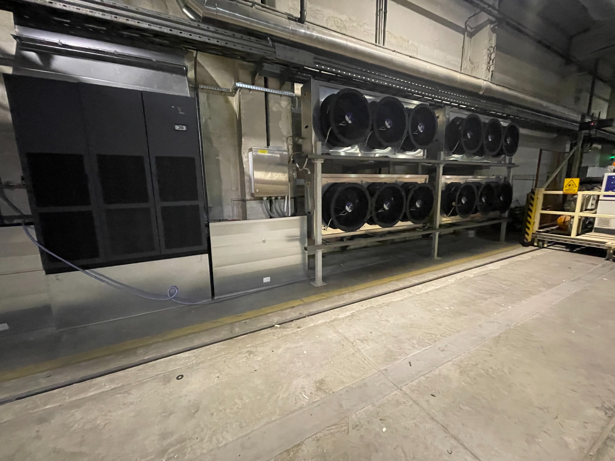 Szafa klimatyzacyjna w przemysłowym magazynie wysokiego składowania, widoczne wentylatory na metalowej konstrukcji i okablowanie.