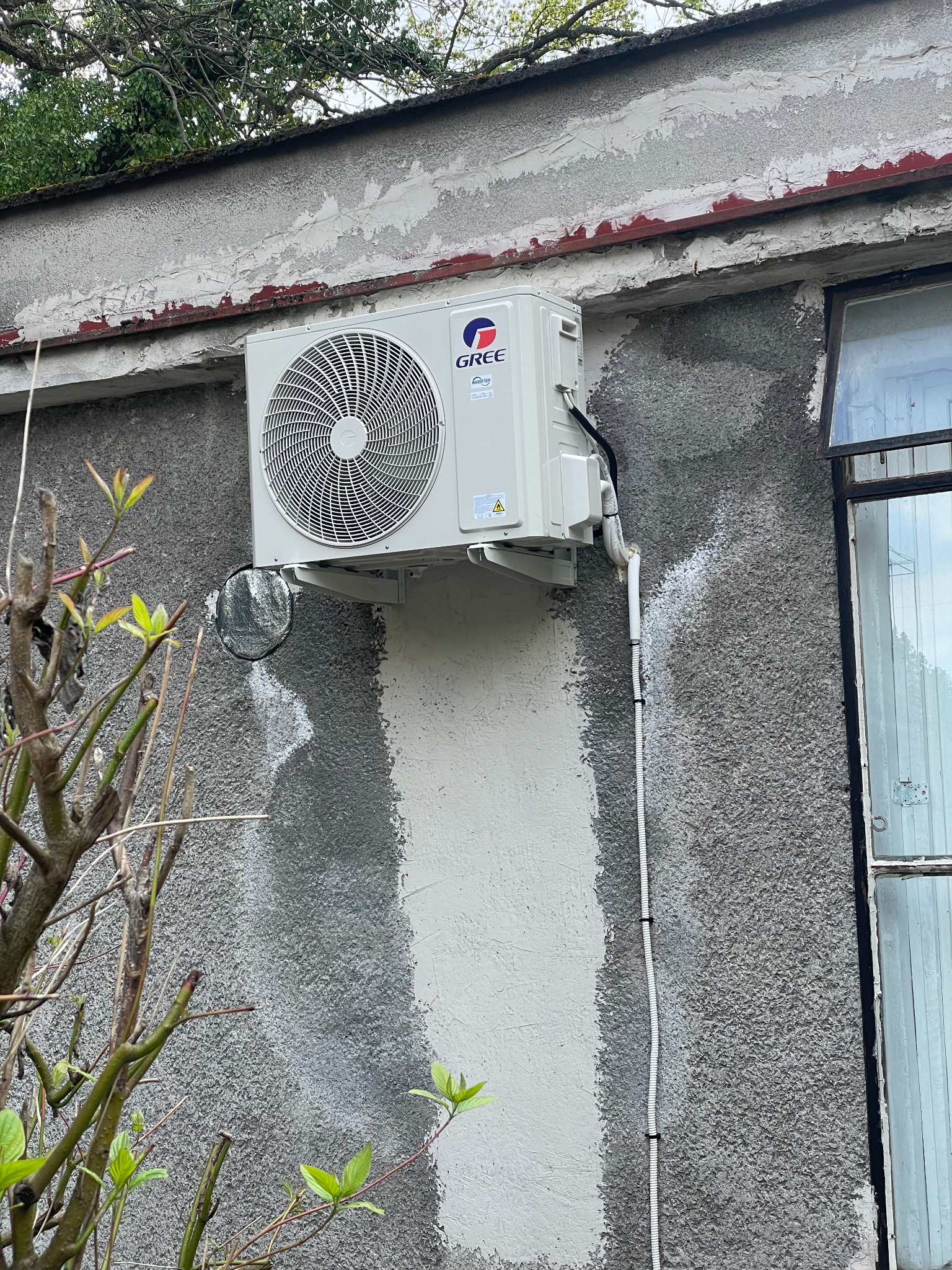 Zewnętrzna jednostka klimatyzatora Gree zamontowana na szarej, chropowatej ścianie budynku z widoczną instalacją odprowadzającą skropliny. Świeżo pomalowany fragment ściany pod klimatyzatorem.