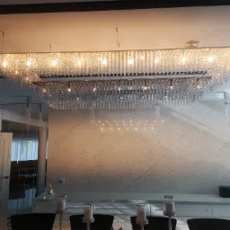 Kryształowy żyrandol o prostokątnej formie, podwieszony nad stołem w jadalni, oświetlający marmurową ścianę i czarne krzesła.