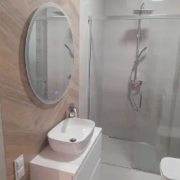 Nowoczesna łazienka z owalnym lustrem z podświetleniem LED, kabiną prysznicową typu walk-in z deszczownicą i umywalką nablatową na białej szafce.