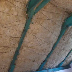 Izolacja poddasza z wełny mineralnej mocowana sznurkiem do pomalowanych na zielono krokwi dachowych.